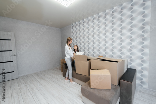 A young mother with children unpacks a box of things after moving to a new apartment. Purchase of real estate. Housewarming, delivery and transportation of goods. © Andrii