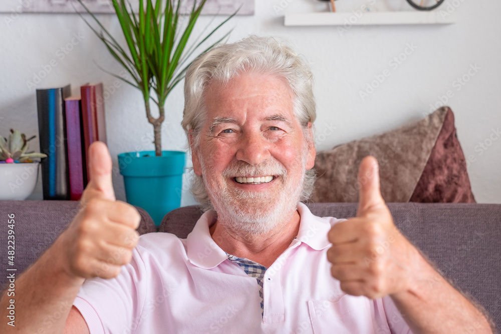 Close up portrait of smiling adult beautiful senior bearded man wearing eyeglasses with both thumbs up, positivity concept. White haired grandfather on sofa at home looking at camera Close up portrait of smiling adult beautiful senior bearded man wearing eyeglasses with both thumbs up, positivity concept. White haired grandfather on sofa at home looking at camera