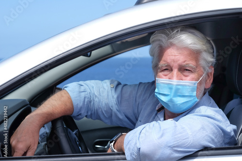 Senior adult man wearing surgical mask due to coronavirus covid 19 omicron variant and delta variant sitting inside the car. Elderly white haired grandparent looking at camera, horizon over sea © luciano