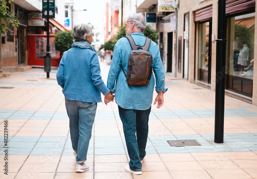 Rear view of active smiling adult Caucasian senior couple walking in the city holding hands as tourists. White-haired retired people dressed in denim enjoying freedom, walking, vacation and travel