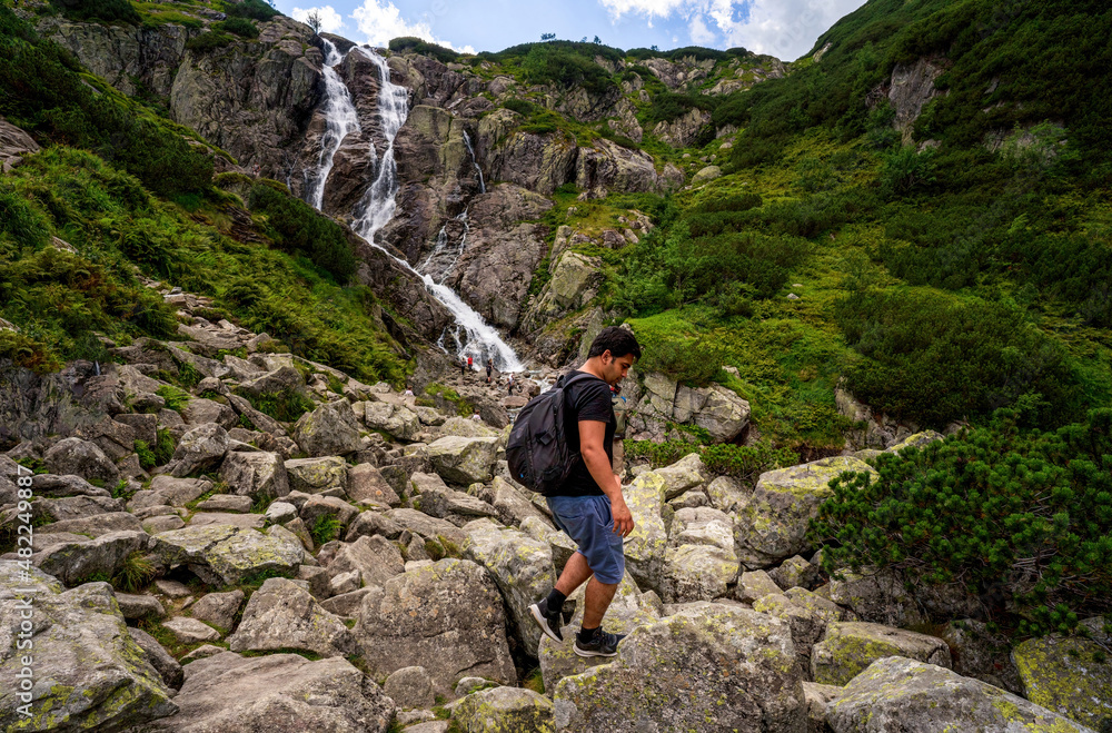 Man hiker tourist with a backpack walks on a rocky mountain terrain, against the backdrop of a huge waterfall in the mountains. Concept of victory, success and achievement of goals