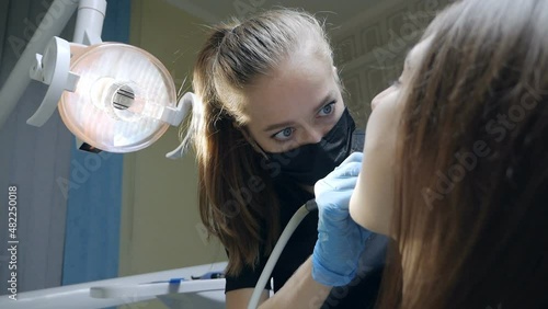 Young woman dentist working with patient's teeth with tools during covid epidemic. The light of the lamp is aimed at the patient's teeth