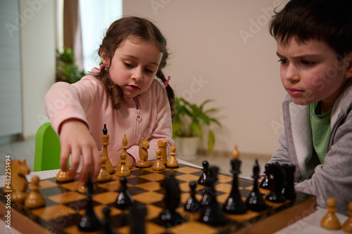 Cute little girl with serious look sitting at the table and plays chess with her brother, picking up a chess piece and making his move. Early development, home educational games for children