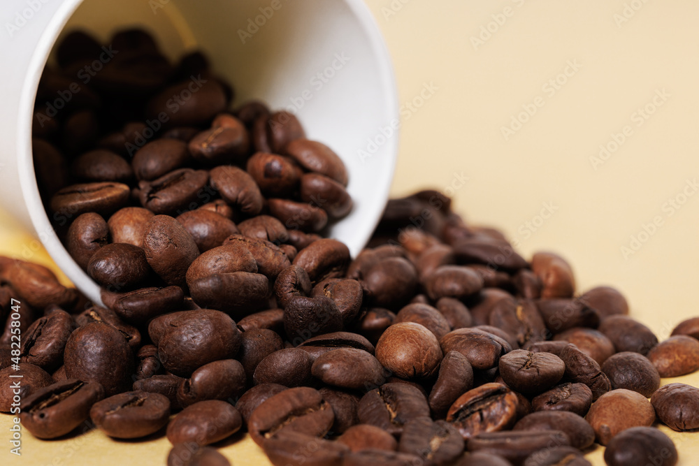 Arabica coffee beans scattered from a white paper cup on a yellow background. Arabica coffee beans scattered from a white paper cup on a yellow background.