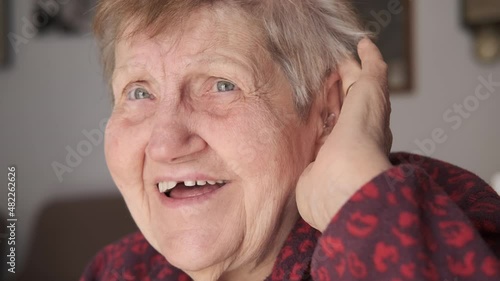 An elderly woman with a hearing aid device in her ear smiles, talks, and tries to listen. One hand on her ear with the device. Senior women with the hearing aid.
 photo