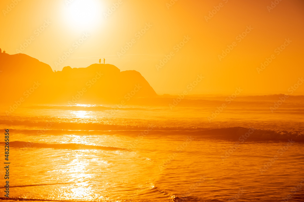 Sunrise on Atlantic ocean with waves and silhouette of rocks with people. Praia Joaquina in Brazil