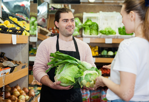 Young male seller offering green onion and cabbage to woman on the supermarket