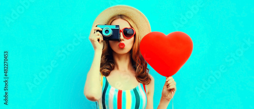 Summer portrait of young woman taking picture on retro film camera with big red heart shaped balloon wearing straw hat on blue background
