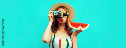 Summer portrait of stylish young woman with retro film camera and slice of fresh watermelon wearing straw hat posing on blue background
