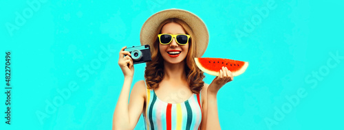 Summer portrait of happy laughing young woman with retro film camera and slice of fresh watermelon wearing straw hat on blue background