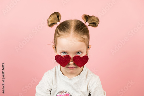 Funny angry little blonde girl in red heart shape sunglasses on pink background. Valentines Day or Mothers Day concept