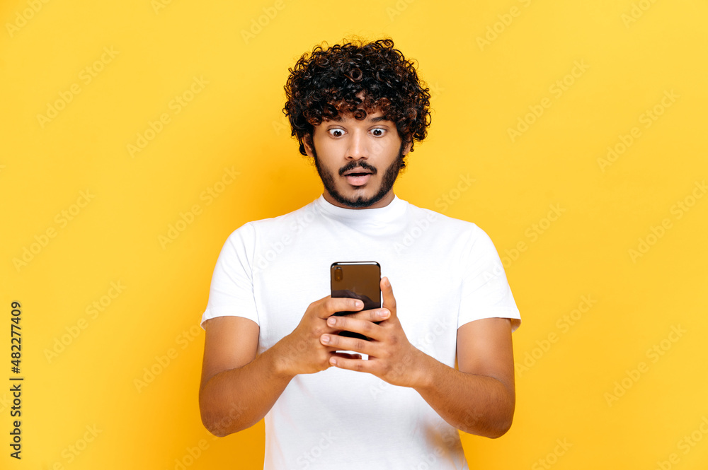 Surprised shocked Indian guy in white t-shirt, uses his smartphone, looks in amazement at screen, received an unexpected message or news, stand on isolated orange background, puzzled facial expression Surprised shocked Indian guy in white t-shirt, uses his smartphone, looks in amazement at screen, received an unexpected message or news, stand on isolated orange background, puzzled facial expression