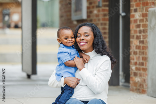 A beautiful African-American mom sitting on steps outdoors and hugging her toddler age son and they are both smiling and happy