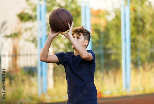 Cute smiling boy in blue t shirt plays basketball on city playground. Active teen enjoying outdoor game with orange ball. Hobby, active lifestyle, sport for kids, teenagers © Natali