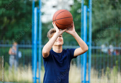 Cute smiling boy in blue t shirt plays basketball on city playground. Active teen enjoying outdoor game with orange ball. Hobby, active lifestyle, sport for kids, teenagers