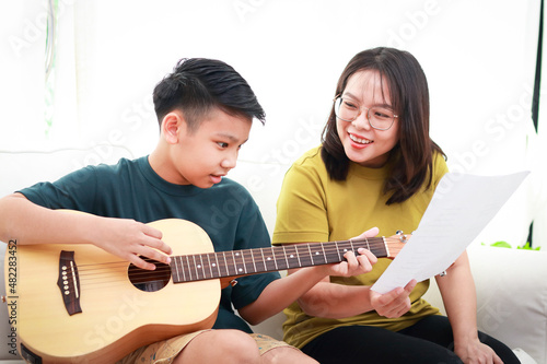 Asian mother and son sitting on the white sofa in the house son plays guitar, mother sings They both enjoyed playing music. Family concept, leisure activities, recreation, international music lessons