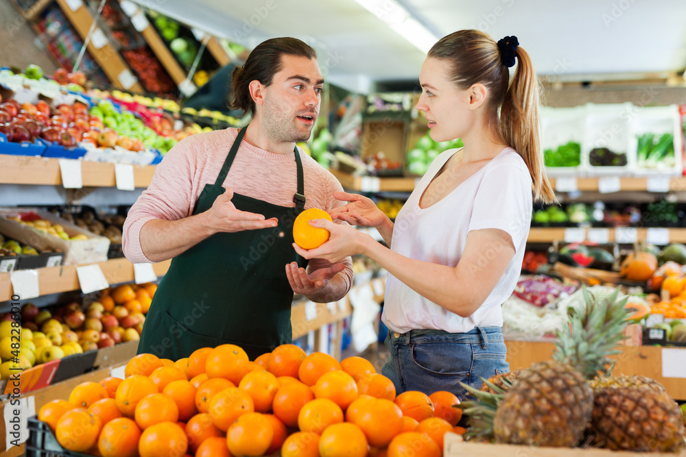 Young man in apron selling fresh oranges to woman customer on the supermarket