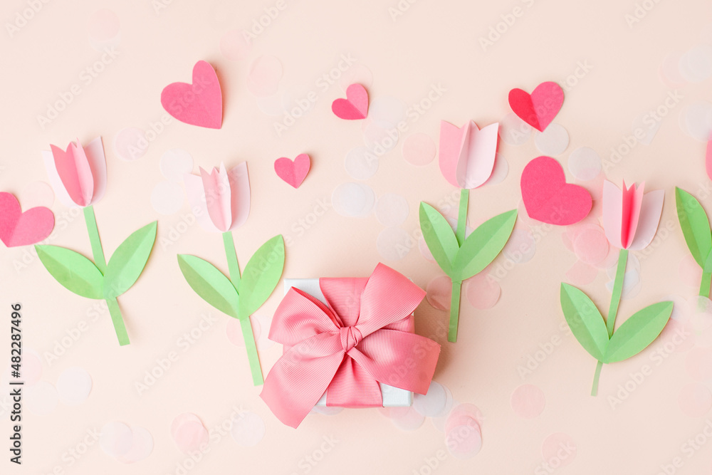 Gift present box with pink bow decorated paper hearts and tulip flowers on pink pastel table top view. Flat lay composition for birthday, valentine, mother day, 8 march, wedding, paper art decor