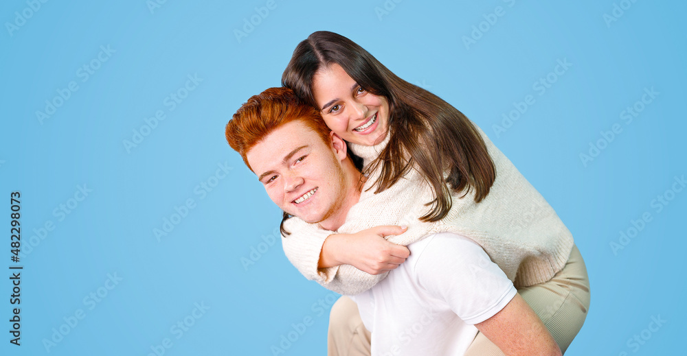 Two young people in love is having fun, pretty joyful lovely smiling girl is sitting on her boyfriend's back isolated on blue background. Two young people in love is having fun, pretty joyful lovely smiling girl is sitting on her boyfriend's back isolated on blue background.
