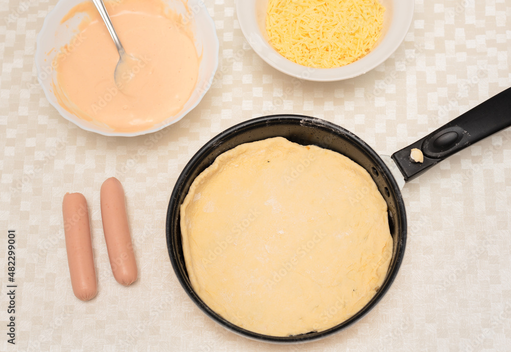 Raw dough on black pan is ready for placing ingredients on it for homemade pizza. Other ingredients are on the table. Top view with selective focus. Process of cooking pizza, step 3