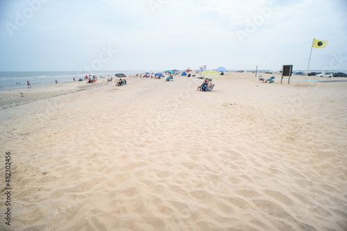 Hot sand under your feet heats up under the summer UV rays over the dunes and onto the seasonal tourist beach in the background, small groups of vacation people sit under sun umbrellas and on chairs photo