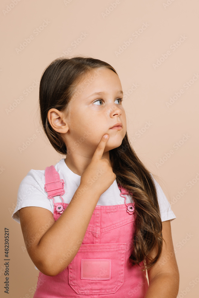 Portrait of child with shining bright eyes looking away, point finger on chin area of face with pensive look wearing bright pink jumpsuit and white t-shirt on beige background. Portrait of child with shining bright eyes looking away, point finger on chin area of face with pensive look wearing bright pink jumpsuit and white t-shirt on beige background.