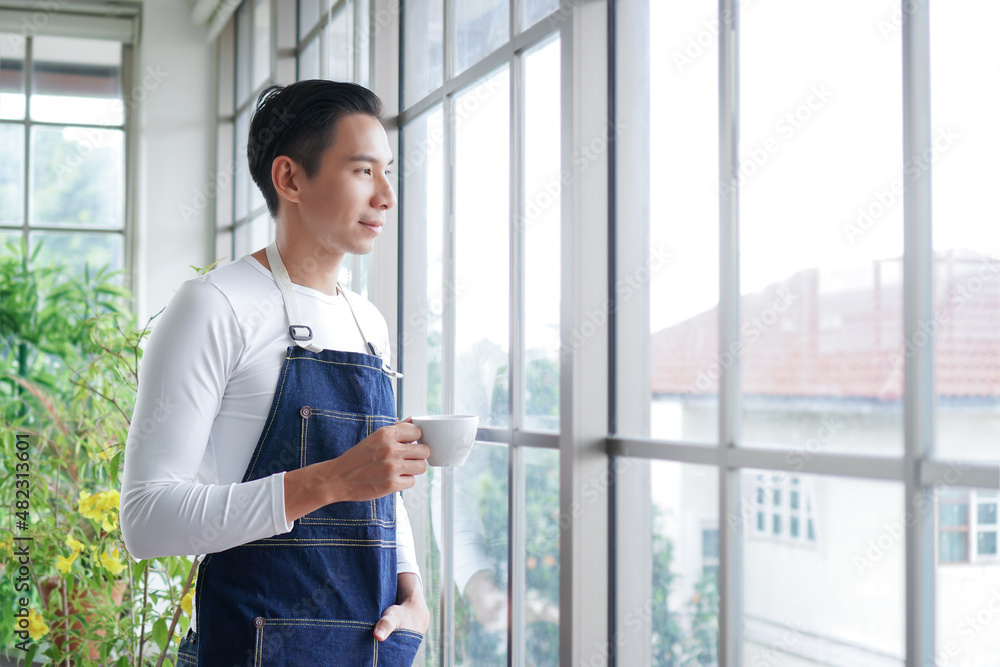 Happy young Asian man wearing apron smiling and holding cup of coffee looking out of window after take care of trees in greenhouse at home Happy young Asian man wearing apron smiling and holding cup of coffee looking out of window after take care of trees in greenhouse at home