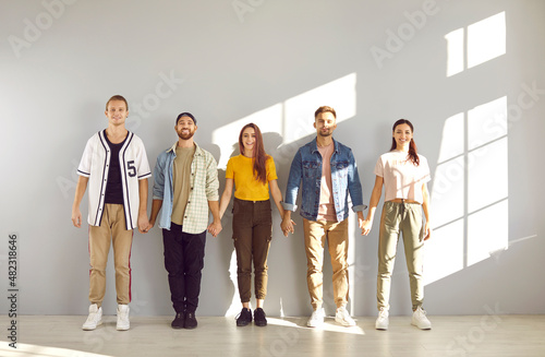 Team of happy young people standing together. Indoor studio group shot of cheerful friends in comfortable informal tees, shirts, pants, trousers and sneakers standing in row, holding hands and smiling