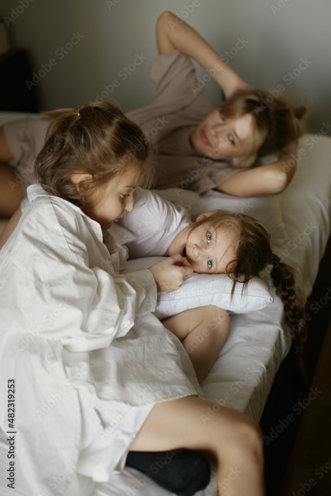 Mother and two funny daughters are laughing and playing together. Sisters. Friendship. Family. Bedroom. Pillows and blanket. Love and happiness. Emotional and cute girls . Casual home clothes. 