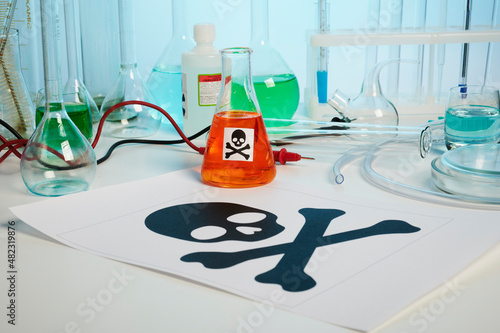 Death printed symbol on a chemical table. Poison fluids in toxicology laboratory. Photo contains danger paper sheet, test tubes, orange colored liquid in chemical flask, some retorts, electrodes photo