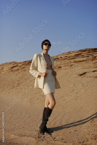 Gorgeous sexy woman walking in the desert. Slim and tan model. Brunette with big lips and long legs. White jacket and skirt, black sunglasses, black boots and turban. Fantastic sand landscape. 