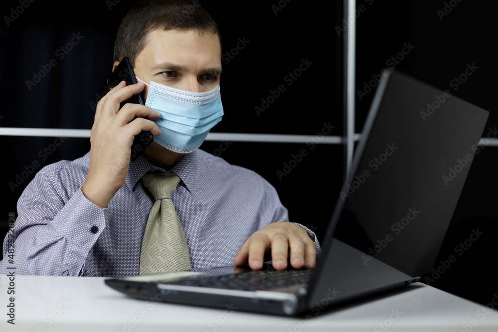Man in face mask talking on mobile phone sitting at laptop. Discussion of working issues, office call, safety during coronavirus pandemic Man in face mask talking on mobile phone sitting at laptop. Discussion of working issues, office call, safety during coronavirus pandemic