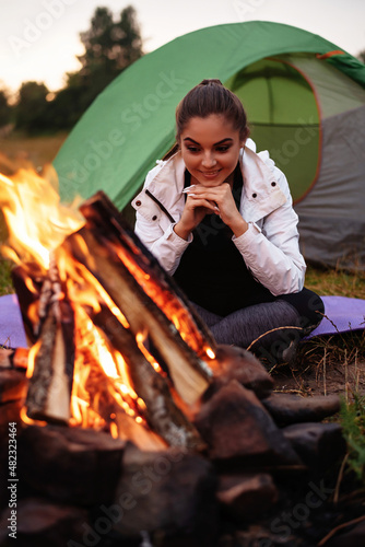 Camping in nature. Beautiful girl on a hike. The girl is warming herself by the fire, smiling and looking to the fire. Green meadow and mountains on the background. Hiking and resting concept. © WellStock