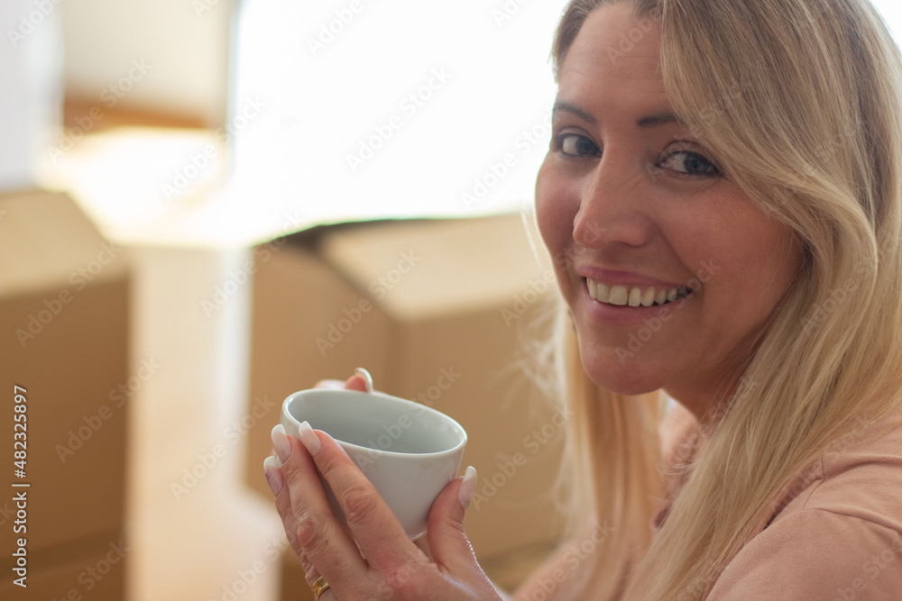 Portrait of relaxed woman in new house. Fair-haired woman in casual clothes sitting, looking at camera smiling, drinking coffee or tea. Real estate, purchase, family concept Portrait of relaxed woman in new house. Fair-haired woman in casual clothes sitting, looking at camera smiling, drinking coffee or tea. Real estate, purchase, family concept