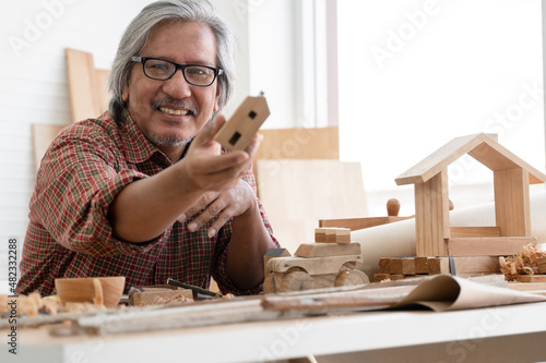 Asian white haired senior carpenter man smiling and showing small wooden house model he made with pride at workplace at home. White background