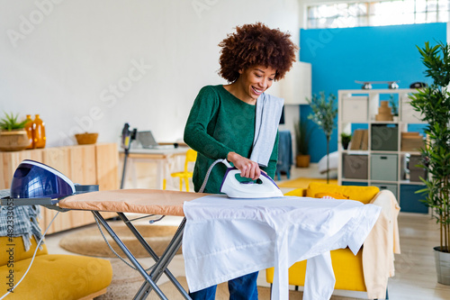 Smiling young Afro woman ironing shirt on board in living room photo