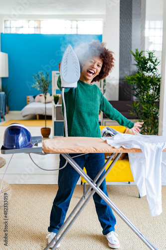 Cheerful young Afro woman blowing steam from iron standing by board with shirt in living room photo