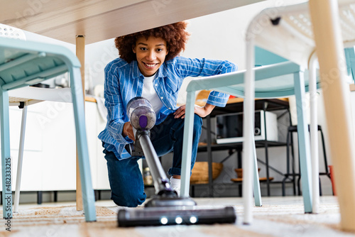 Smiling young woman crouching using vacuum cleaner under dining table at home photo