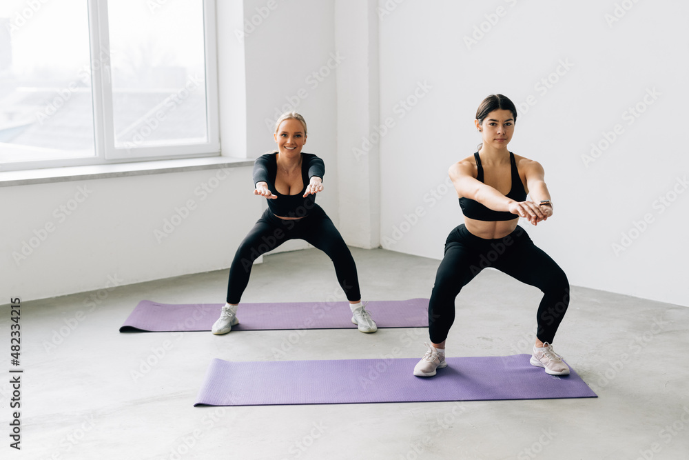 Tough stamina training for two young women stunning fitness models doing squats together indoors. Tough stamina training for two young women stunning fitness models doing squats together indoors.
