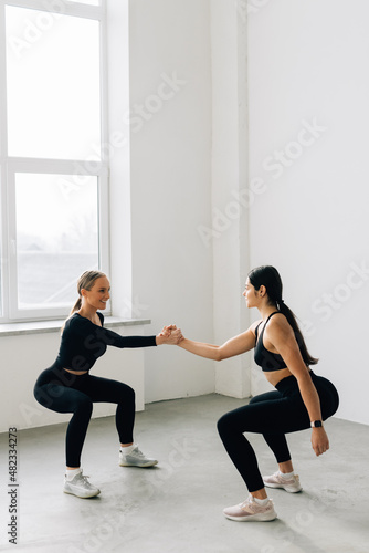 Fitness pose two sporty women, blonde and brunette, in black sportswear show squats in studio, isolated at white, help each other holding hands - healthy lifestyle, training and friendship concept