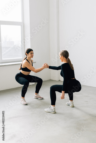 Fitness pose two sporty women, blonde and brunette, in black sportswear show squats in studio, isolated at white, help each other holding hands - healthy lifestyle, training and friendship concept
