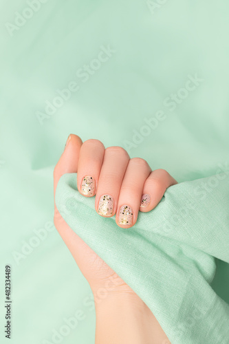 Female hand with clear nail design. Glitter clear nail polish manicure with green nail art. Woman hand hold light green fabric on green background.