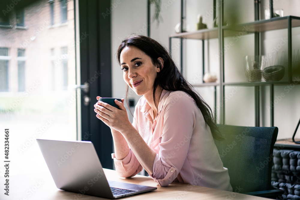 Smiling businesswoman with laptop having coffee at desk in coworking office Smiling businesswoman with laptop having coffee at desk in coworking office