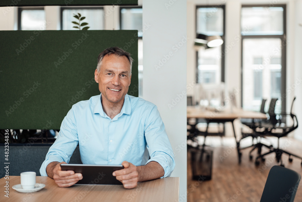 Smiling mature businessman with tablet PC sitting at desk in coworking office Smiling mature businessman with tablet PC sitting at desk in coworking office