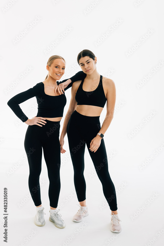 Two concentrated women dressed in sports wear bonding to each other while standing indoor against white background. People, exercising and sport concept Two concentrated women dressed in sports wear bonding to each other while standing indoor against white background. People, exercising and sport concept