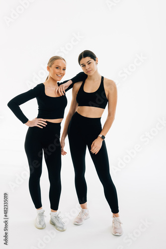 Two concentrated women dressed in sports wear bonding to each other while standing indoor against white background. People, exercising and sport concept
