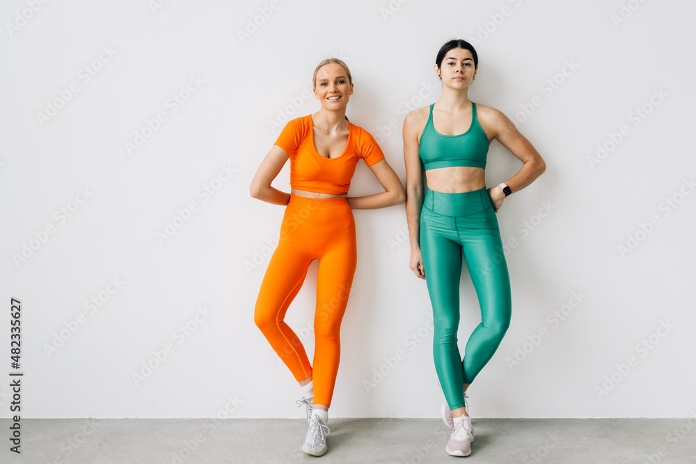 Fototapeta premium Two sporty blonde and brunette girls in athletic body cloth sport wear cloth stand together after workout on white background