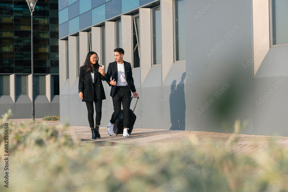 Businessman with trolley bag walking with colleague on footpath by office building Businessman with trolley bag walking with colleague on footpath by office building
