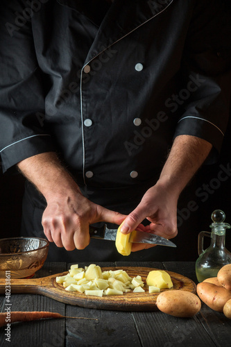 The chef cuts raw potatoes into pieces with a knife before preparing breakfast or dinner. Close-up of a cook hands while working in a restaurant kitchen.