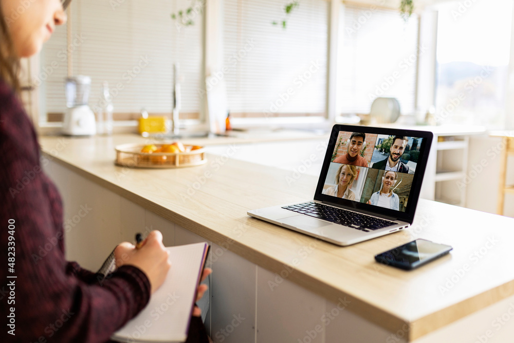 © Westend61 - Young working woman with notebook doing video call with colleagues on laptop at home